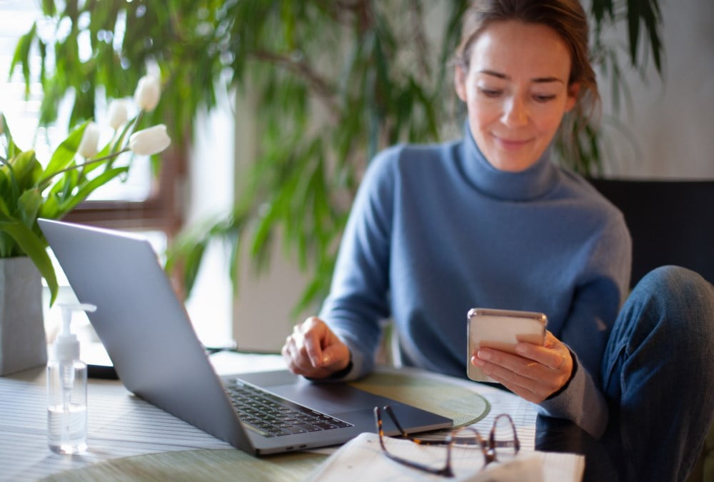 Ung kvinne sitter komfortabelt ved et hjemmekontorbord omgitt av grønne planter og jobber på en laptop mens hun holder en smarttelefon. Hun smiler mens hun ser på telefonen, med briller og en notatbok ved siden av seg på bordet.