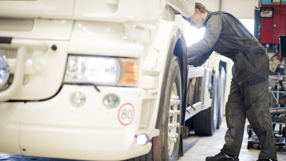 Mekaniker i grå arbeidsklær lener seg på lasteplanet til en hvit lastebil.